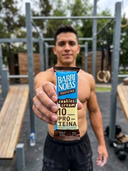 A close-up, first-person perspective shot of a fitness enthusiast holding a branded protein bar in a gym environment.
